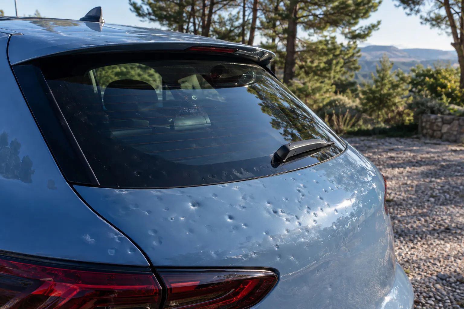 Coffre et lunette arrière d'une compacte bleu clair marqués de petits impacts de grêle, sur un parking de campagne.
